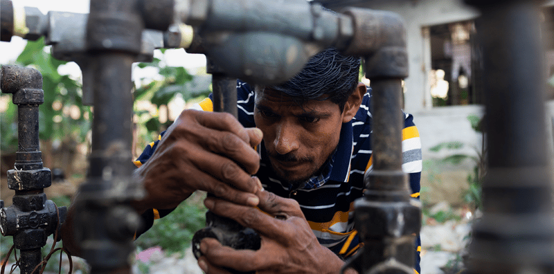 A man in a striped shirt works on rusted industrial pipes, using both hands to adjust a valve in a close-up outdoor setting with greenery in the background.
