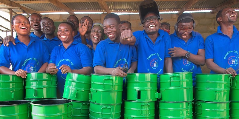 Group of people in matching blue shirts smiling and laughing behind bright green metal buckets, raised hands and hands-on-hips in a lively, celebratory scene.