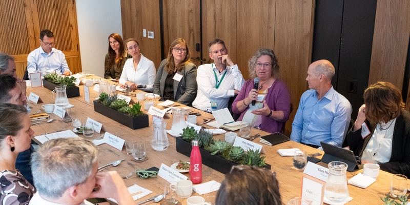 A diverse group of professionals sits around a long wooden table in a conference room, one person speaking with a microphone while others listen attentively.