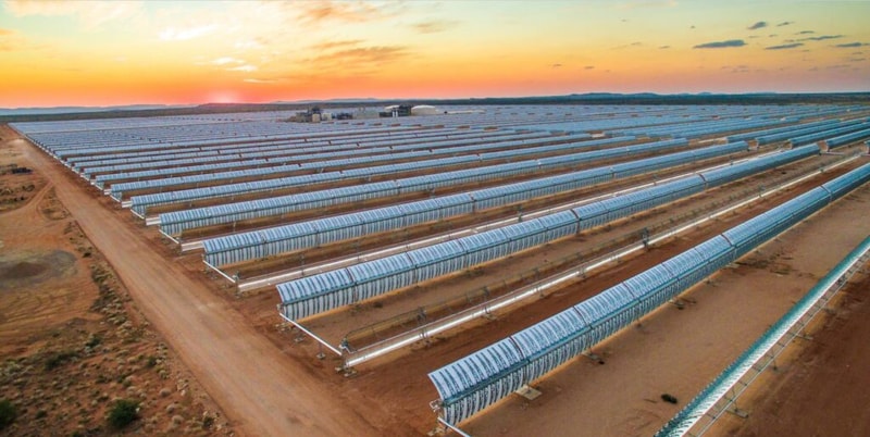 Rows of greenhouse-like glass structures in a desert landscape at sunset, with long, parallel covers stretching across the dry terrain.