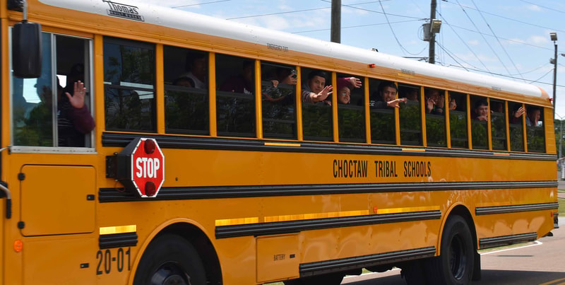 Yellow school bus with students waving from windows, "CHOCTAW TRIBAL SCHOOLS" lettering, stop sign extended, parked on a sunny day with power lines in background.