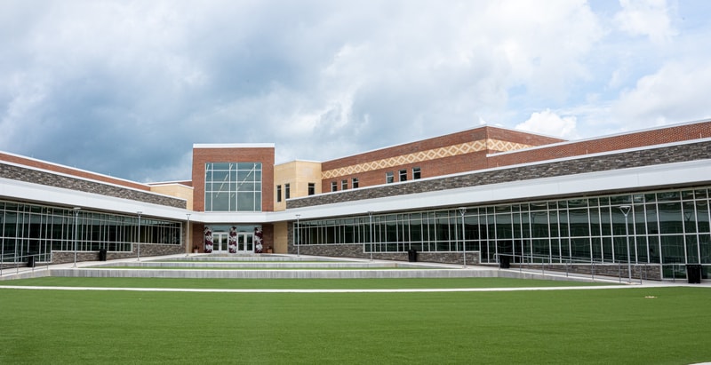 A modern brick-and-glass campus building with large windows, decorative patterns, and a wide grassy lawn in the foreground under a cloudy sky.