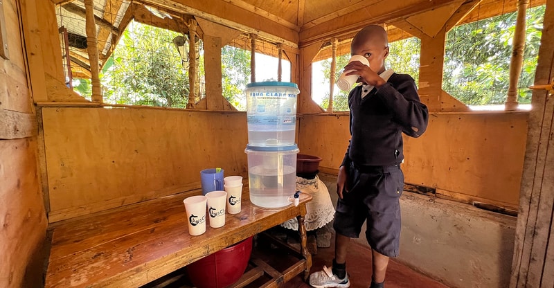 A person stands in a wooden shelter drinking from a white cup beside a tall water dispenser; several cups on a bench and a blue pitcher nearby.