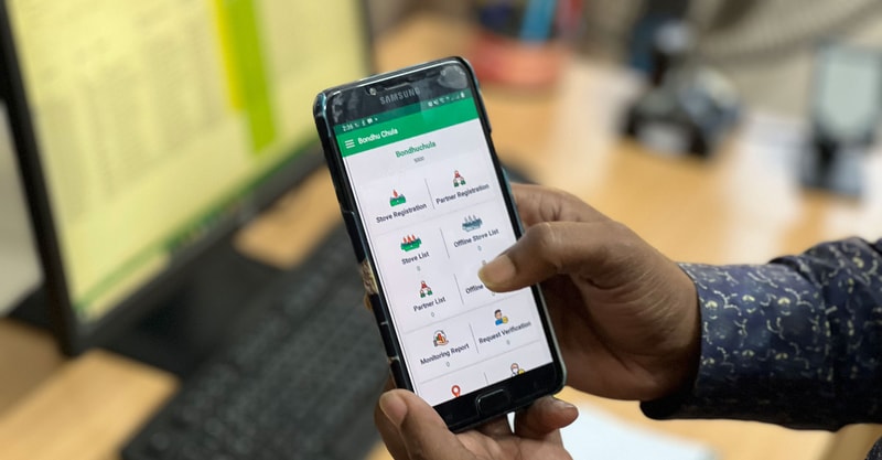 A hand holding a Samsung smartphone displaying a green-themed app with icons and labels such as Stove Registration, Partner Registration, Store List, and Online Stove List.