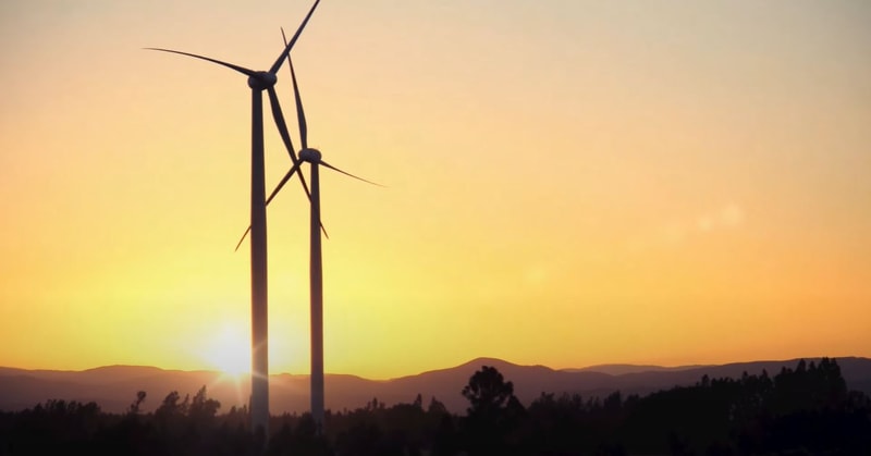 Two tall wind turbines stand silhouetted against a bright sunset, with a warm orange sky, distant hills, and a dark treeline near the horizon.