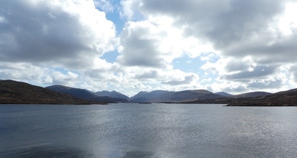 A wide calm lake stretches to distant hills under a partly cloudy sky, with light reflections on the water and rugged, softly rolling mountains on the horizon.