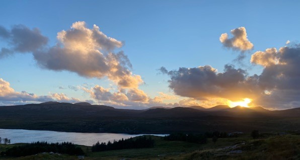 Golden sunset over rolling hills with a calm lake in the foreground, scattered fluffy clouds illuminated in warm orange hues against a clear blue sky.