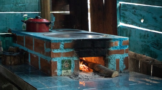 Traditional brick outdoor oven with a metal stovetop, blue-washed walls, and a crackling wood fire visible inside the firebox; a red kettle sits on the left.