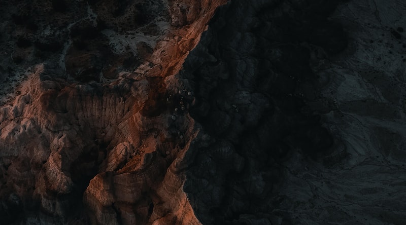 Aerial view of rugged rocky cliff edges with warm orange-brown sunlit rock on left transitioning to dark shadowed terrain on the right, revealing jagged textures and crevices.