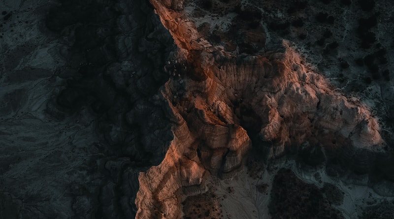 Aerial view of dark rocky cliff faces with a central reddish-orange rock formation illuminated, jagged textures, shadows, and rugged desert terrain.