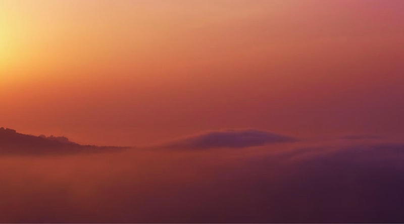 Soft, warm sunrise or sunset sky with gradient orange to pink hues; indistinct distant hills shrouded in low-lying, hazy fog across the horizon.