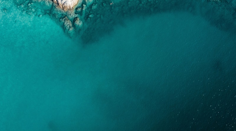 Aerial view of turquoise ocean with a rocky shoreline along the top left, transitioning to deep blue water on the right.