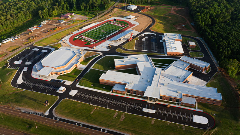 Aerial view of a modern school campus with a large football stadium, surrounding classrooms and gym buildings, parking lots, and surrounding wooded area.