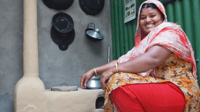 Smiling woman in colorful traditional attire sits beside a clay stove with pots hanging on a gray wall and a green fence in the background.
