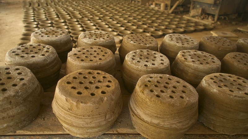Stacks of traditional clay pot lids with multiple small holes, drying in a dusty workshop with more lids arranged in rows in the background.