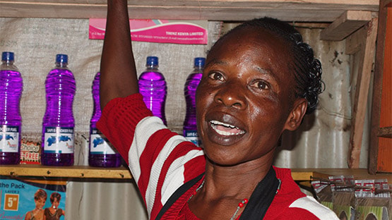 A woman with braided hair in a red and white striped shirt stands behind a shelf of purple bottles, raising one arm and speaking toward the camera.