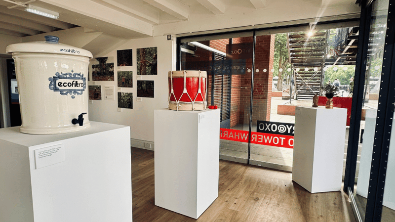 A bright exhibition space with white pedestals displaying a large ceramic water dispenser, a red drum, and small crafted items, glass doors and wooden floor visible.