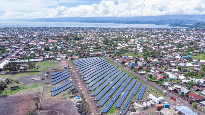 Aerial view of a town with a large solar farm feature: long rows of solar panels on open fields, surrounded by houses, greenery, and a distant coastline.