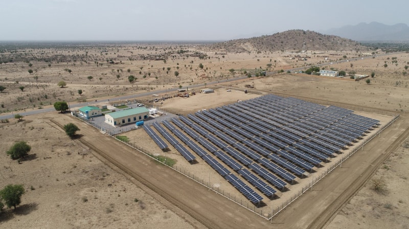 Aerial view of a solar farm with many solar panels in neat rows, adjacent buildings, a dirt road, and a dry desert landscape with sparse trees and distant hills.