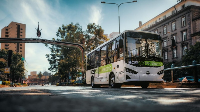 Electric white city bus with green “100% electric” decals drives down a sunny urban street under an arched gateway, with buildings and trees lining the road.