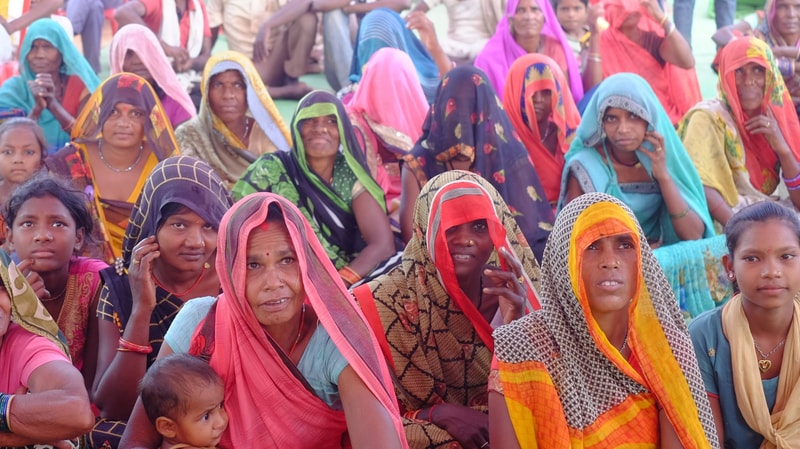 A large group of Indian women and children sit together outdoors, many wearing colorful sarees and headscarves, looking toward the camera.