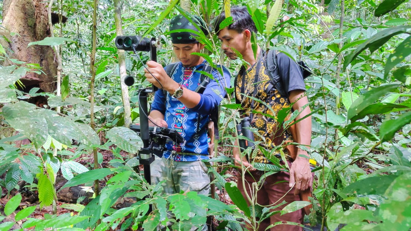 Two people in a dense jungle setting operate a mounted camera or recording rig, with one person adjusting the device while the other watches, both wearing patterned shirts and backpacks.