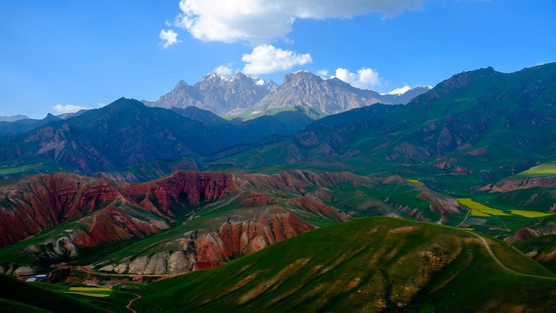 Vast mountainous landscape with deep green hills, red rock formations in the foreground, terraced fields, and snow-capped peaks under a bright blue sky with scattered clouds.