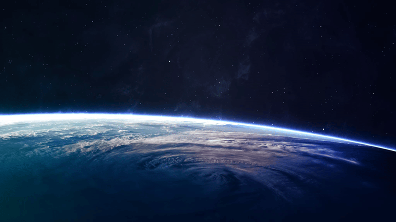 An image of Earth’s curved horizon from space, showing a thin bright blue atmosphere edge above dark starry space with swirling cloud patterns across the globe.