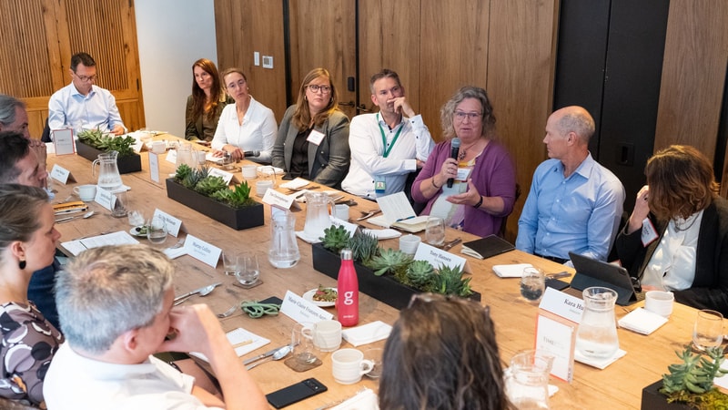 Group of professionals seated around a long wooden table in a meeting room, one woman speaking into a microphone while others listen attentively. Name cards, notebooks, and plants decorate the table.