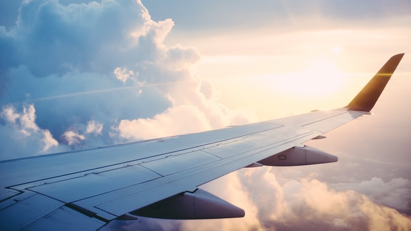 Airplane wing extending across a cloudy sky with sunlight breaking through, soft blue and orange hues from a sunset or sunrise.