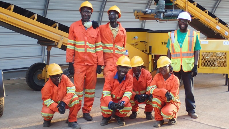 Group of construction workers in orange safety gear and yellow helmets posing in front of yellow machinery inside an industrial site.