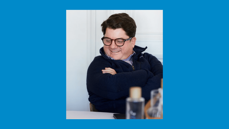 Man smiling with arms crossed, wearing glasses and a dark navy zip-up sweater, seated indoors at a table with bottles and a phone in view.