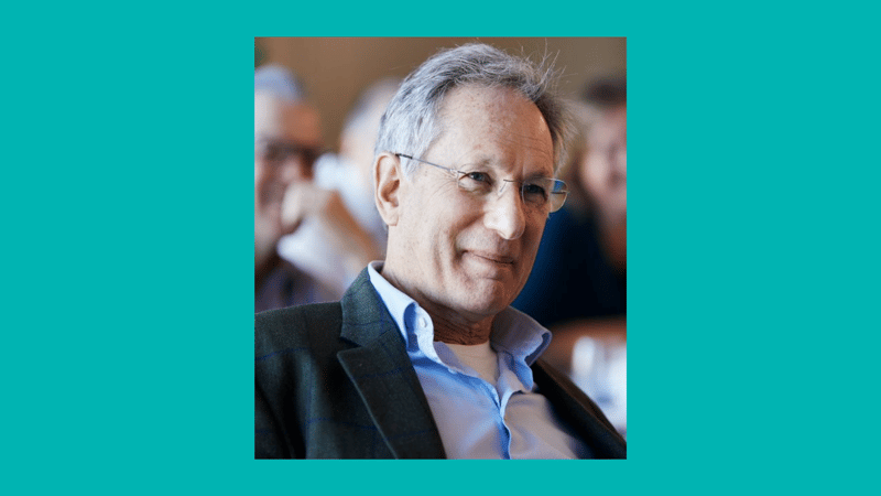 An older man with gray hair and glasses, wearing a blue shirt and dark blazer, smiling slightly in a candid setting with blurred people in the background.