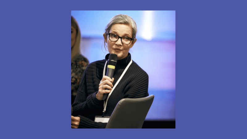 woman with short gray hair and glasses speaks into a microphone on stage, wearing a black ribbed sweater and a white lanyard, seated with a chair in foreground