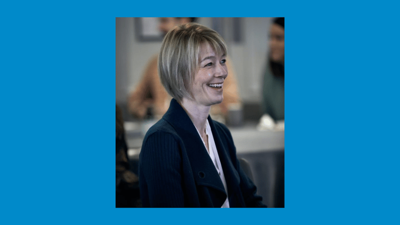 Smiling woman with short blonde hair wearing a dark blazer in a professional setting, candidly engaged and laughing, with blurred colleagues in the background.