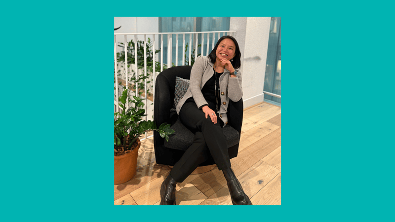 A woman sits in a dark armchair on a wooden floor, wearing a gray cardigan, black top and pants, black ankle boots, smiling with one hand resting on her chin.