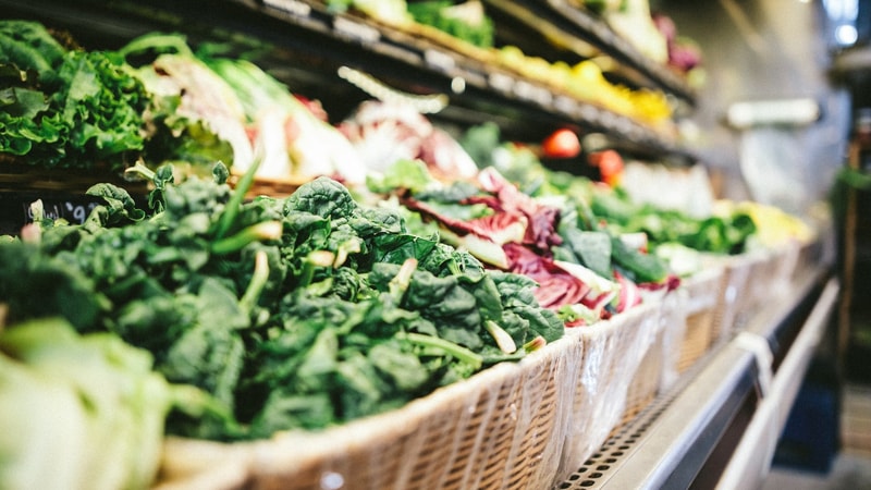 A close-up of fresh leafy greens and colorful lettuce arranged in a market basket on a grocery store produce aisle, with shelves and other vegetables in the blurred background.