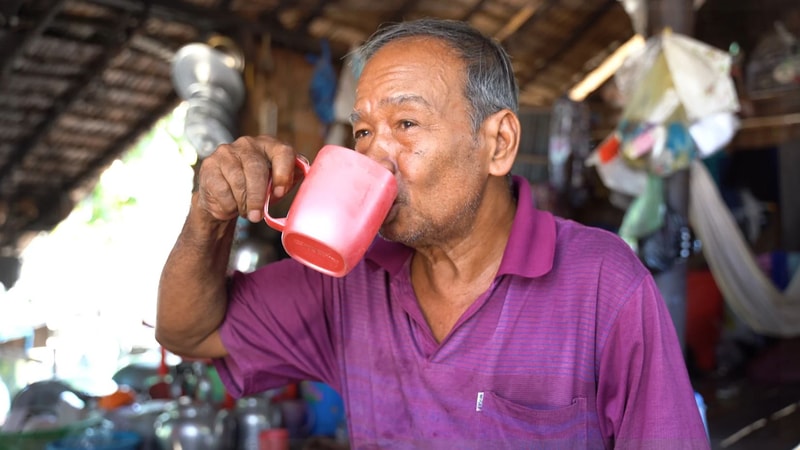 An older man with short gray hair wearing a purple shirt is drinking from a pink mug inside a rustic, cluttered indoor space.