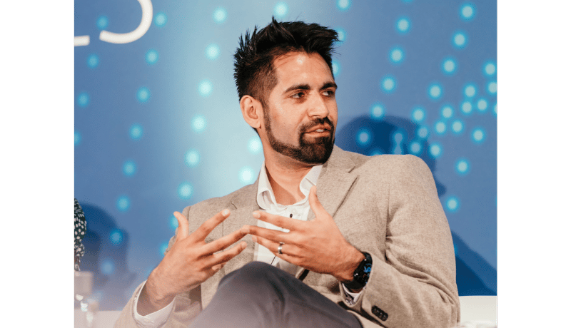 Man with dark hair and beard wearing a light tan blazer and white shirt speaks, gesturing with both hands in conversation on a panel stage with blue bokeh backdrop.
