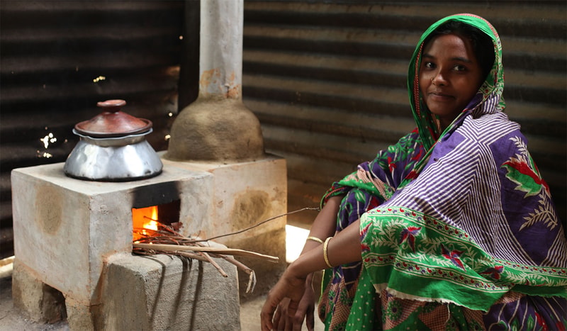 Woman in a colorful sari sits beside a small brick stove with a fire, a metal kettle on top, in a simple rustic setting.