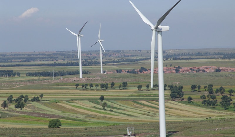 Southwestern field landscape with three tall white wind turbines across rolling green farmland, small trees, scattered buildings in the distance under a blue sky.