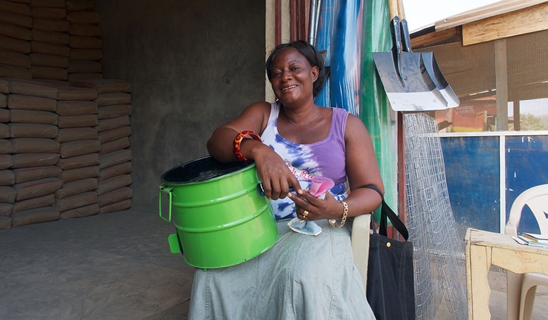 A smiling woman sits on a chair in a workshop, holding a bright green metal bucket and pink cloth, with tools and a bag nearby. Bags of cement stacked in background.