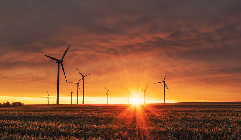 Row of wind turbines across a flat field at sunset, golden light bursting from the horizon, long shadows, dramatic orange and purple sky.