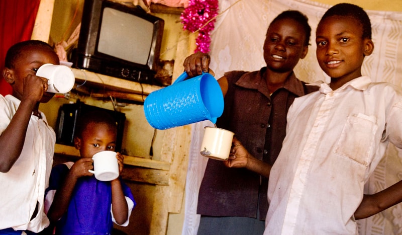 Children and two adults indoors share a cheerful moment as a blue bucket pours liquid into a beige mug, while others sip from white cups.