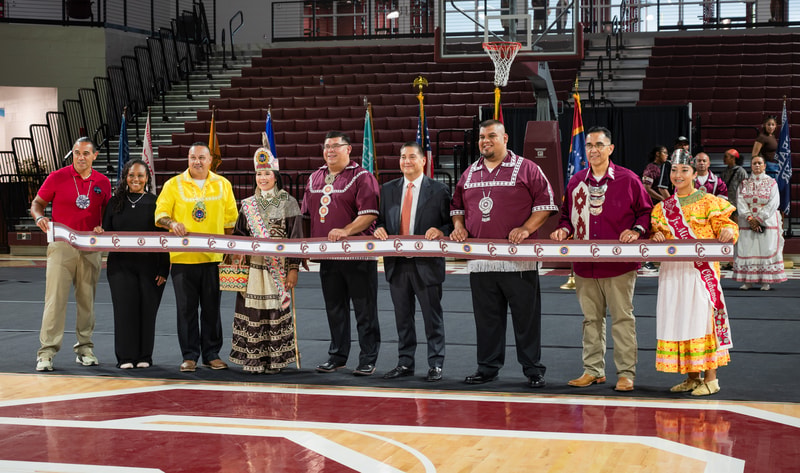 Group of diverse people in ceremonial attire standing together in a gymnasium, holding a long ribbon for a grand opening or ribbon-cutting event.