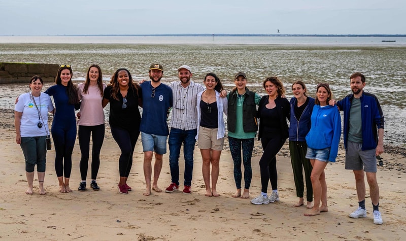 group of fourteen people standing arm-in-arm on a sandy beach, smiling, with the sea and sky in the background.