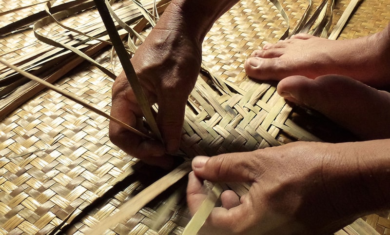 Hands weaving dry palm fronds into a woven mat, threading and shaping narrow strips over a textured, straw-colored surface.