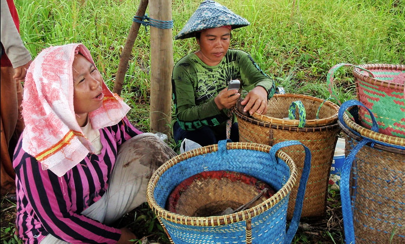 Two people resting in a grassy area with woven baskets; one wears a pink striped shirt with a towel on their head, the other in a green shirt with a blue hat, using a mobile phone.