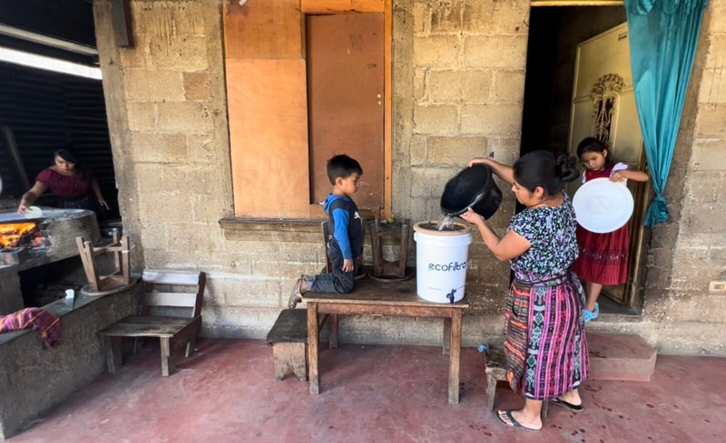 A woman pours water from a pot into a large white bucket labeled “eco filtro” while a child sits on a wooden bench and another child stands in a doorway holding a plate.