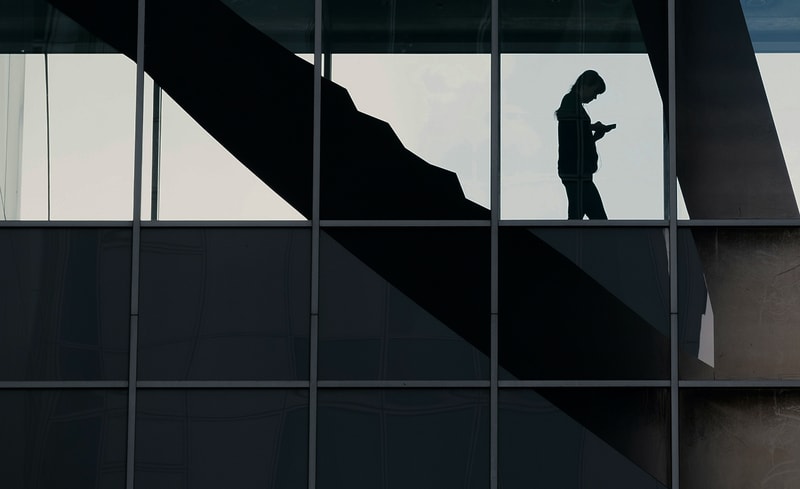 A silhouette of a person standing on an upper-floor glass-walled staircase inside a modern building, seen through large window panes with angled structural shadows.